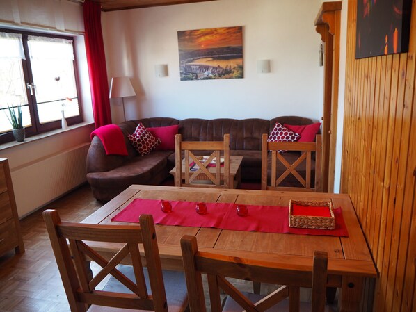 Un salon chaleureux avec un canapé marron, des coussins rouges, une table en bois sur laquelle est posée une nappe rouge, et un tableau représentant un coucher de soleil accroché au mur.