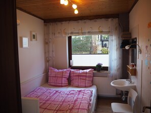 Une petite chambre à coucher avec un lit, une fenêtre avec des rideaux, un radiateur, un lavabo et un miroir.