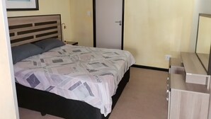 Un dormitorio con cama, mesita de noche, cómoda, puerta y un cuadro enmarcado en la pared.