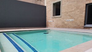 Una piscina con zona poco profunda de 1,2 metros, rodeada por un muro de ladrillo y un edificio de color oscuro.