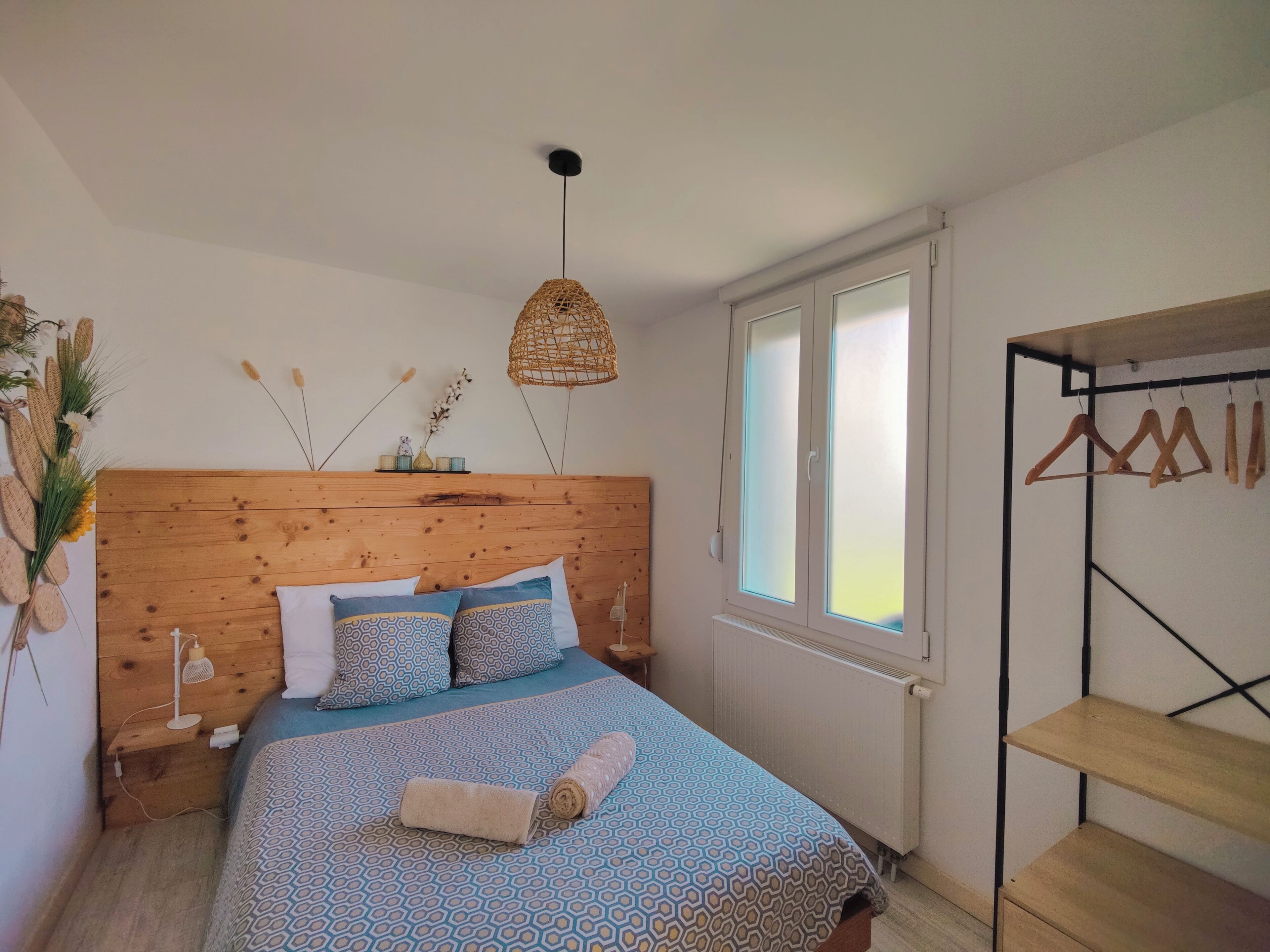 Un dormitorio con cabecera de madera, ropa de cama estampada, lámpara de noche, lámpara colgante, ventana con persianas y un perchero.