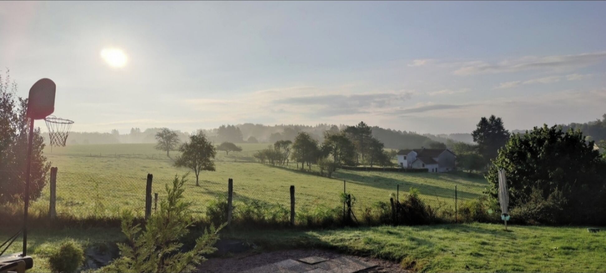 Paisaje rural con un aro de baloncesto, campos abiertos, árboles dispersos y una casa distante.