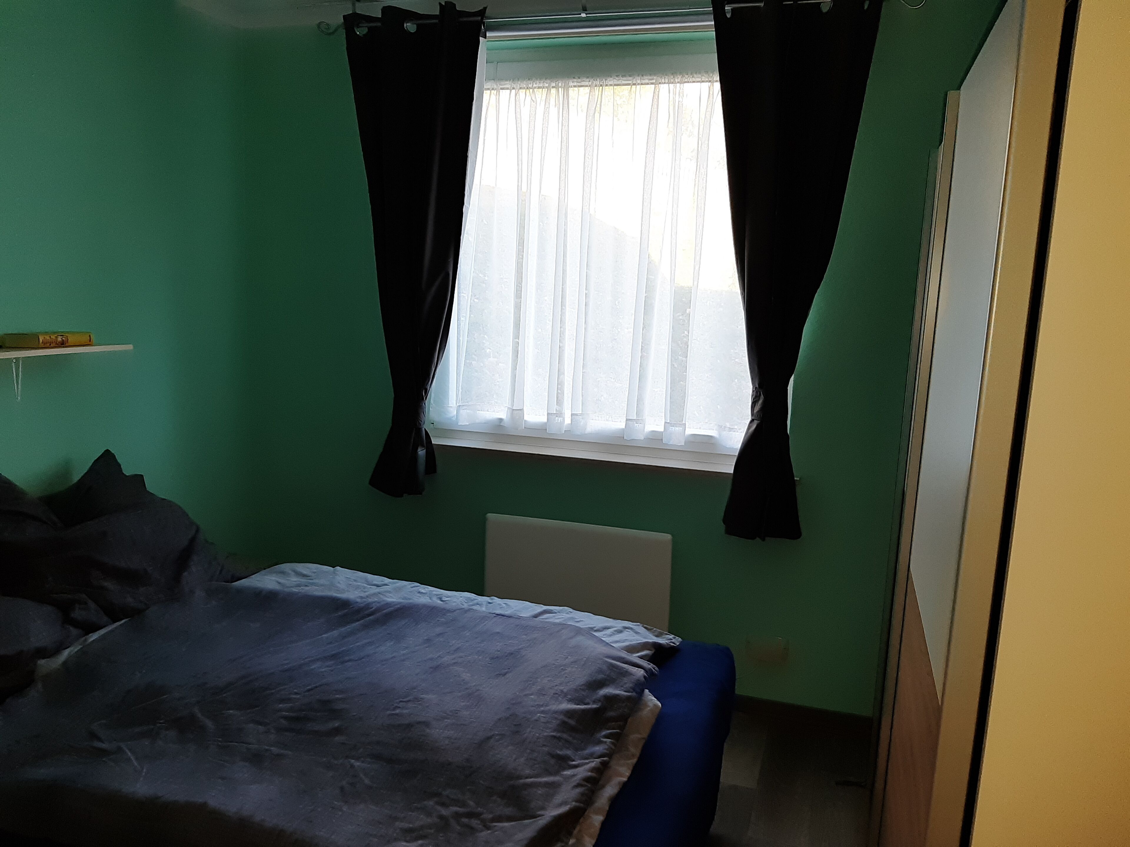 A room with a bed, a window with curtains, a radiator, and a shelf with a yellow object.