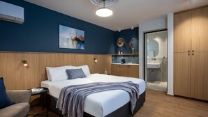 A modern bedroom with a large bed, bedside tables, a chair, a small table with books, a painting on the wall, and a bathroom with a sink and mirror.