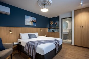 Un dormitorio moderno con una cama grande, mesitas de noche, una silla, una pequeña mesa con libros, un cuadro en la pared y un baño con lavabo y espejo.