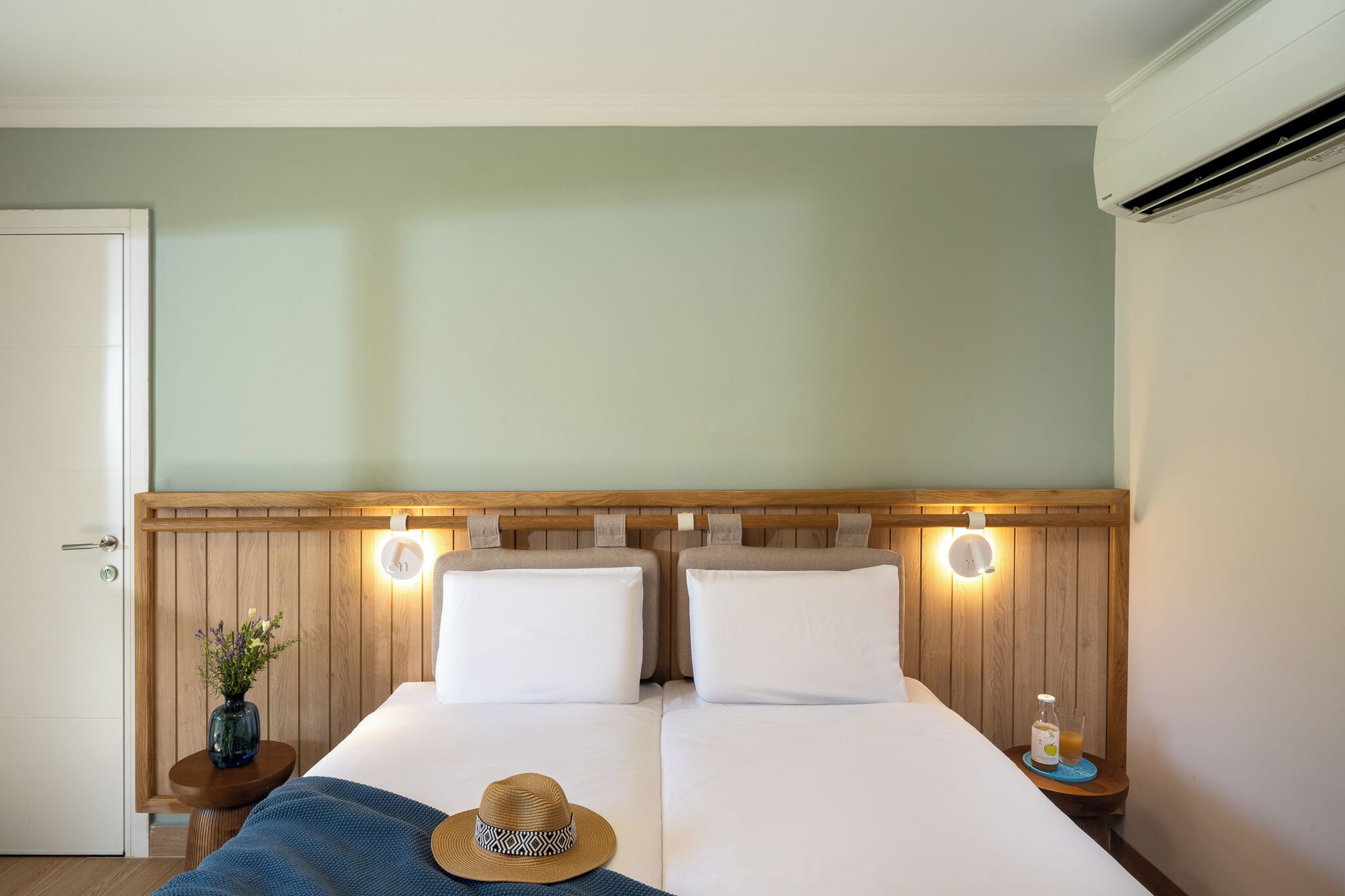Una cama doble con dos almohadas, cabecera de madera, lámparas de noche, un florero con flores, un sombrero y una botella de jugo sobre una mesita de noche.