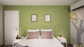 A bedroom with two beds, a white door, two framed pictures on the wall, a small table with a plant, and a straw hat on the wall.