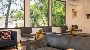 A modern living room with a grey sofa, a wooden coffee table with a book, a bottle of orange juice, and a straw hat. There are two armchairs and a small table with a plant. Large windows offer a view of trees and a building outside.