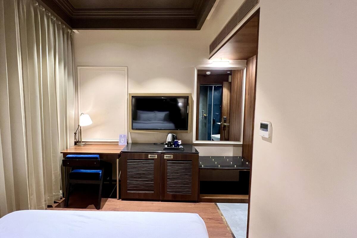 A hotel room with a bed, a desk with a lamp, a TV, a minibar, and a mirror.