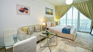 A modern hotel room with a sofa, a bed, a glass coffee table, and a view of the city through large windows. - Gld - Cozy Studio with balcony (Dubai)