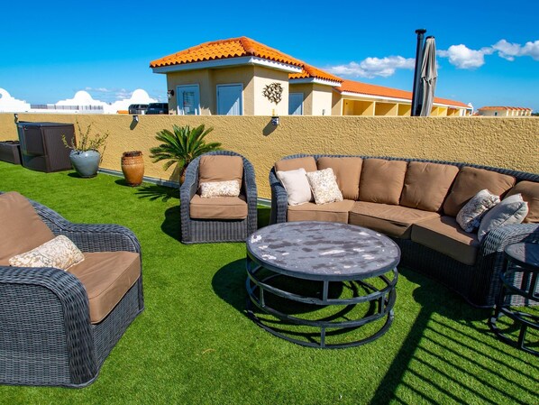 An outdoor patio with wicker furniture, a round table, and a view of a building with a red roof. - Paradise Cove Townhome 145-3 - Premium Waterfront Community (Clearwater Beach)