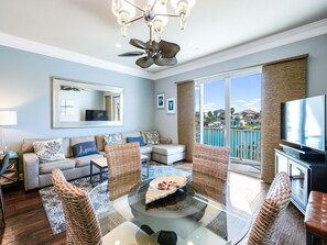 A modern living room with a glass coffee table, wicker chairs, a sofa with patterned cushions, a large mirror, and a television. - Paradise Cove Townhome 145-3 - Premium Waterfront Community (Clearwater Beach)
