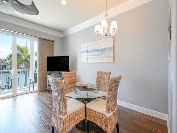 A dining area with wicker chairs, a glass table, a flat-screen TV, and a painting on the wall. - Paradise Cove Townhome 145-3 - Premium Waterfront Community (Clearwater Beach)