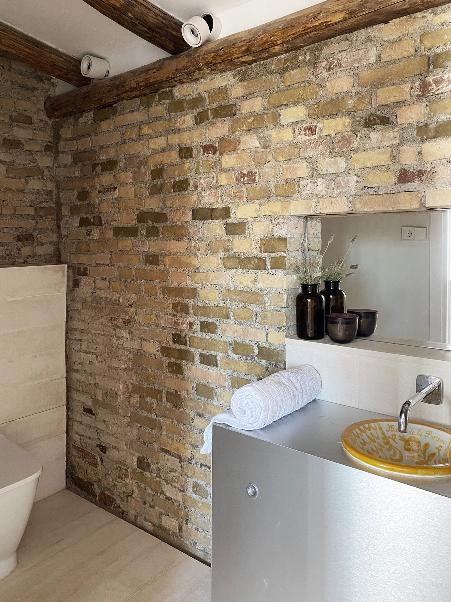 Een badkamer met een bakstenen muur, een wit toilet, een wastafel met een gele kom en een spiegel boven de wastafel.