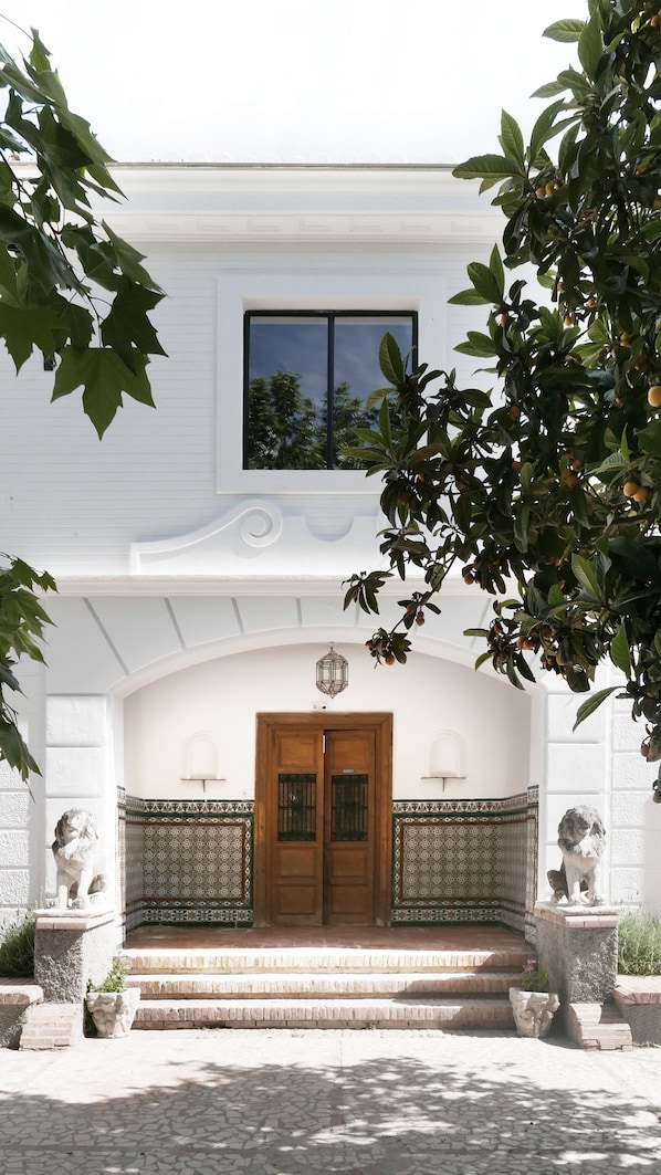 A white building with a wooden double door, a small window above the door, and a decorative balcony railing. - Orchard of the Purisima (Santa Fe)