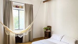 A bedroom with a bed, a hammock, a window with curtains, a radiator, a small table with a plant, and a bookshelf. - Orchard of the Purisima (Santa Fe)