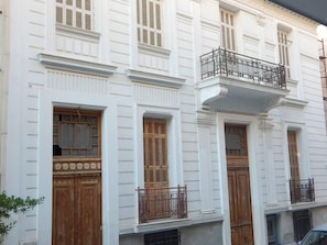 A white building with wooden doors and windows, a balcony with a wrought iron railing, and a small garden area.