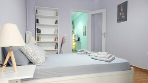 Een slaapkamer met een bed, nachtkastje, lamp, boekenplank en een deur naar een andere kamer.