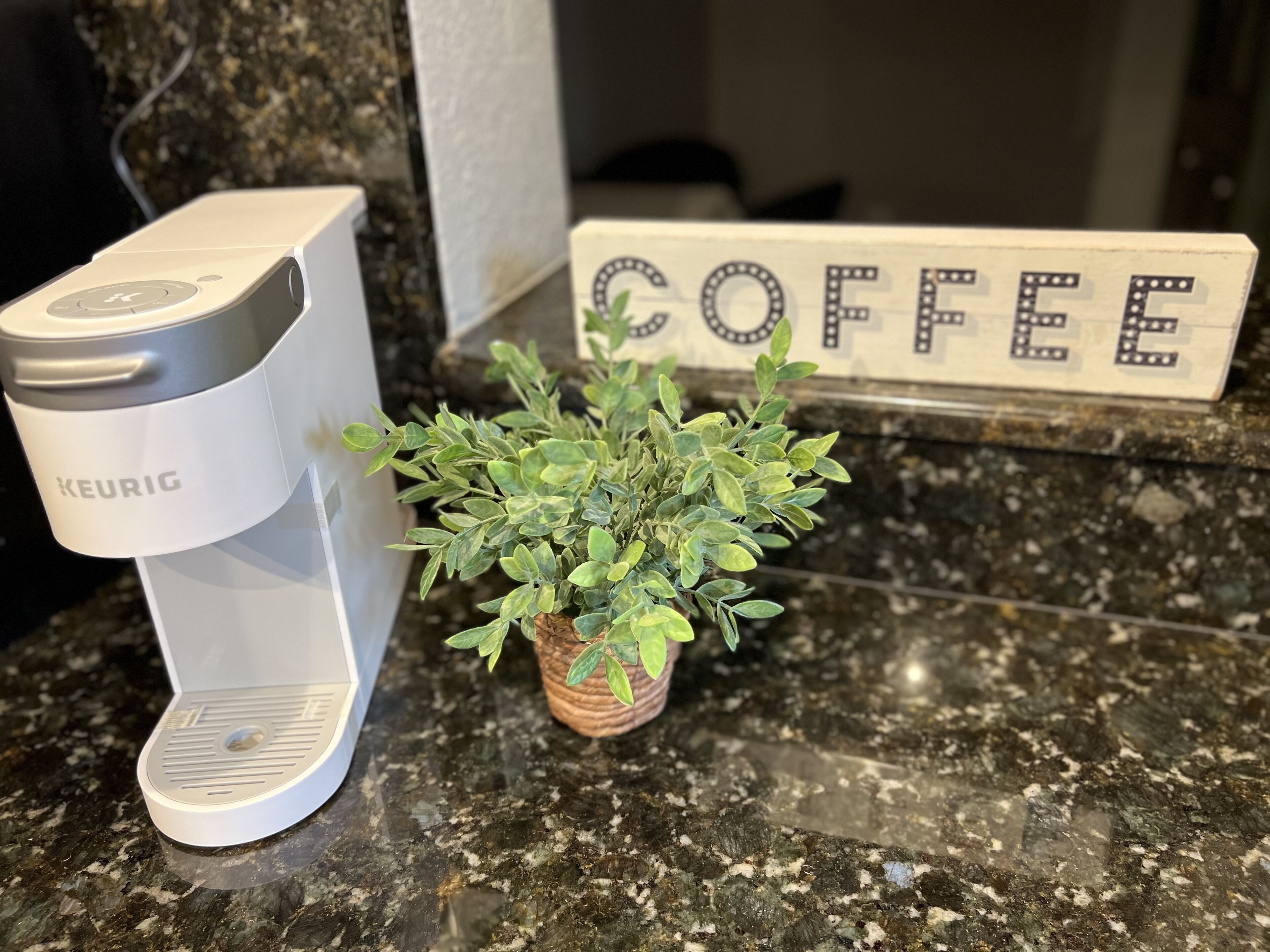 Une machine à café Keurig posée sur un comptoir en granit, avec une petite plante en pot et un panneau indiquant « CAFÉ ».