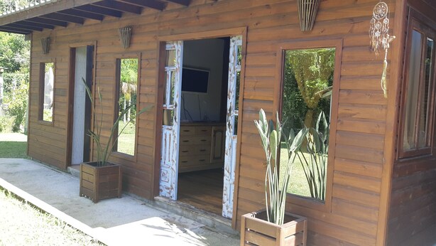 A wooden cabin with open doors revealing a living area, a small TV, and potted plants. - Ibiraquera Lagoon Studio (Imbituba)