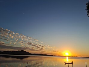 A calm lake with a sunset, a silhouette of a mountain range, and a small boat. - Ibiraquera Lagoon Studio (Imbituba)