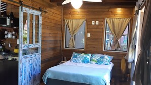 Een houten kamer met een bed, een raam met gordijnen, een keukenhoek met een fornuis en een spoelbak, en een deur met decoratief ontwerp.