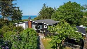 Ett modernt hus med ett svart tak, vita väggar och en skorsten. Det är omgivet av frodig grönska och har utsikt över havet.