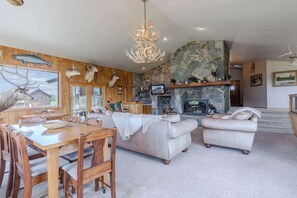 A spacious living room with a stone fireplace, a dining area with wooden furniture, and a large chandelier. - Otter Lodge (West Yellowstone)
