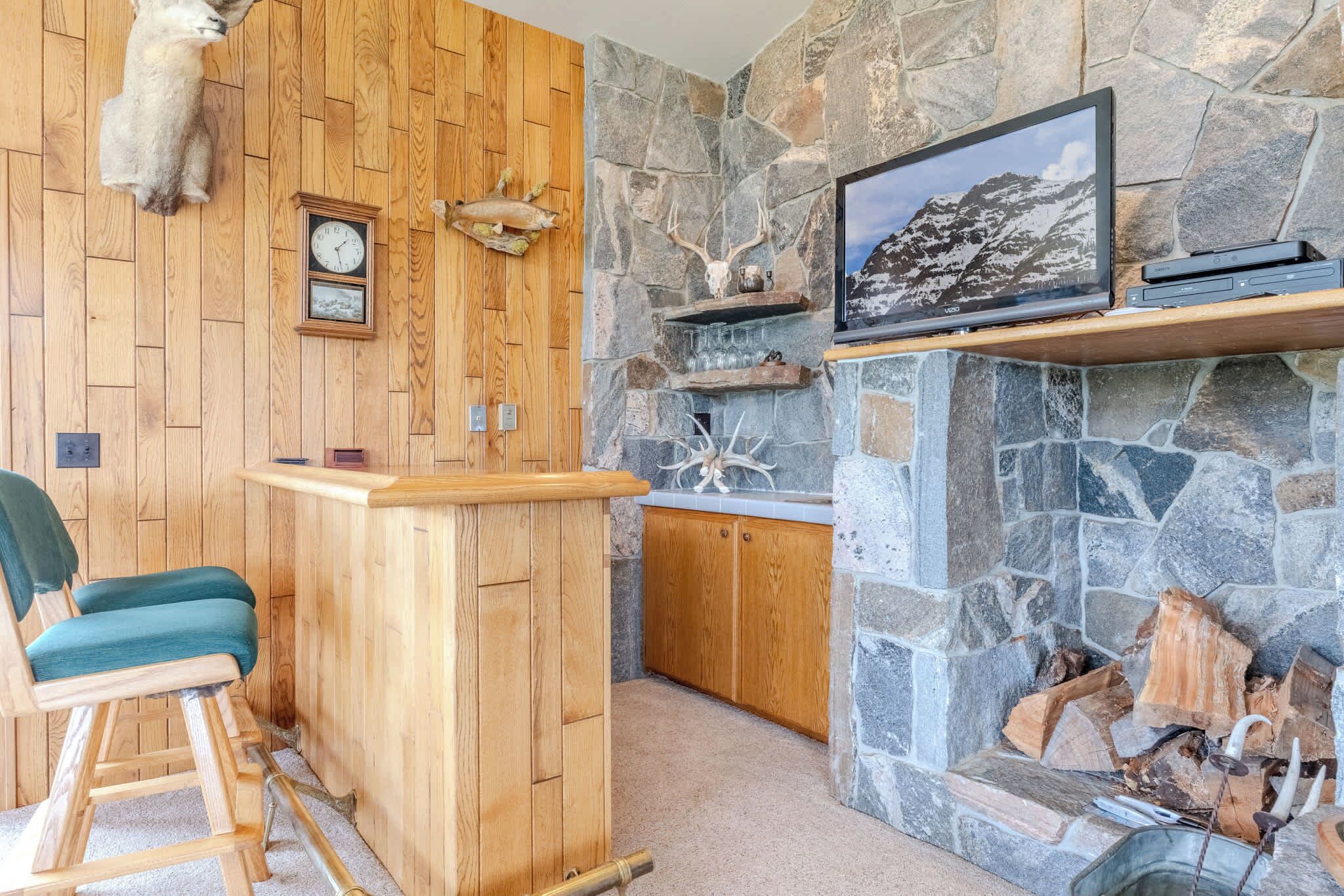 A wooden bar area with a stone fireplace, a mounted deer head, and a framed picture on the wall.