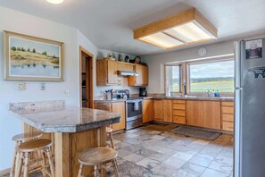 A kitchen with wooden cabinets, a granite countertop, a stainless steel refrigerator, and a window with a view of the countryside. - Otter Lodge (West Yellowstone)