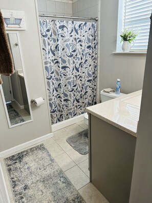 A bathroom with a shower curtain, a toilet, a sink, a mirror, a towel rack, a rug, and a window with a plant. - Guest suite (Chattanooga)