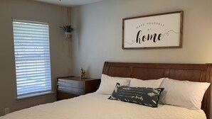 A bedroom with a bed, a ceiling fan, a window with blinds, a bedside table with a plant, and a wall with a motivational quote.