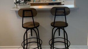 A compact kitchen with a white countertop, two bar stools, wooden cabinets, a window with blinds, and a ceiling light.