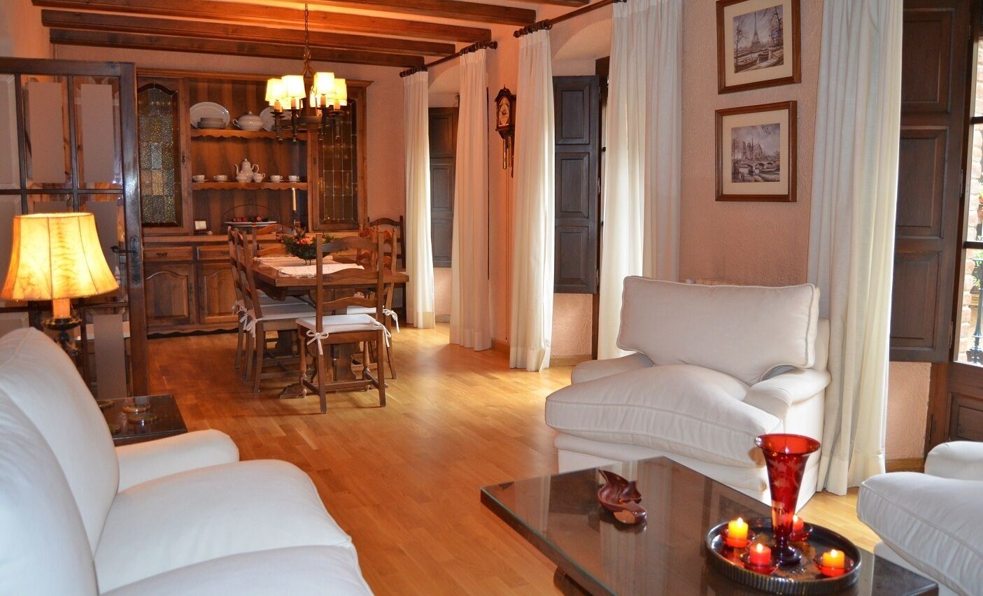 A living room with a wooden floor, white sofas, a dining area with a table and chairs, and a red vase on a coffee table.