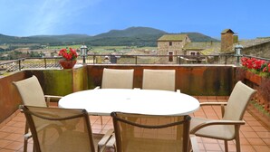 Una terrazza con sedie in vimini e un tavolo, con vista su un paesaggio suggestivo che include colline e un edificio storico.