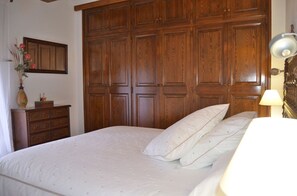 A bedroom with a large wooden wardrobe, a bed with white bedding, and a small dresser with a vase of flowers.