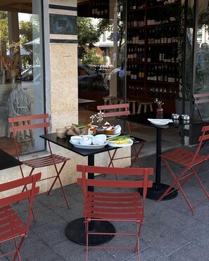 Un área de comedor al aire libre con sillas y mesas de metal rojo, una estantería para vinos al fondo y vista a la calle.
