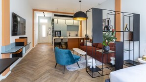 Een modern appartement met keuken, eethoek en woonkamer. Er is een blauwe stoel, een bank en een aan de muur bevestigde televisie.