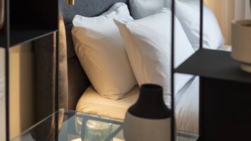 Una cama bien tendida con sábanas y almohadas blancas, una mesita de noche con superficie de cristal y un florero blanco.