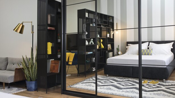 Een moderne slaapkamer met een bed, een grote zwarte boekenkast, een grijze bank en een potplant.