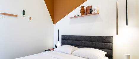 A modern bedroom with a bed, bedside tables, a nightstand, and a wall-mounted shelf with decorative items.