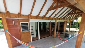 Un espace extérieur abrité, doté d’une structure en bois, d’un hamac et offrant une vue sur un espace intérieur comprenant un coin repas et des chaises.