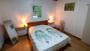 Une chambre avec un plafond en bois, un lit recouvert d’une couvre-lit à motifs floraux, deux lampes de chevet, une fenêtre avec des stores et un fauteuil.