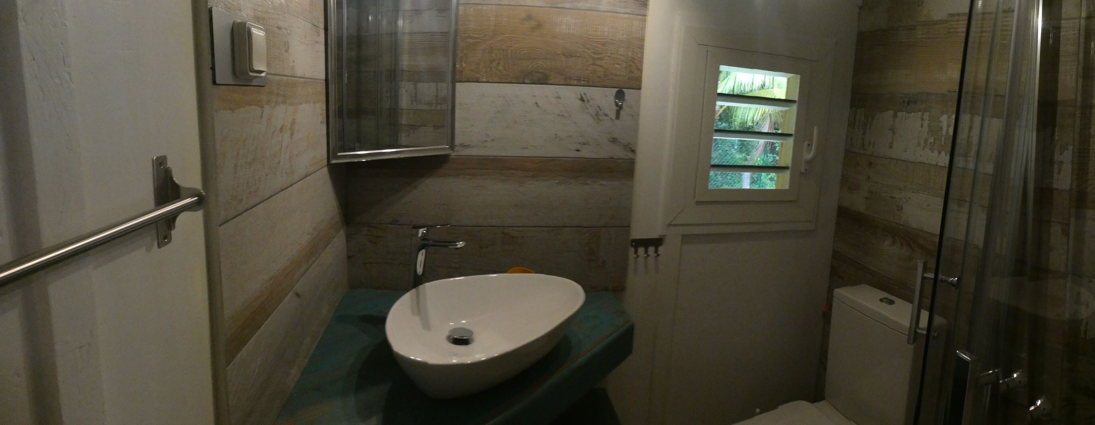 Ein Badezimmer mit einem weißen Waschbecken, einer Duschkabine aus Glas und einem kleinen Fenster mit Blick ins Grüne.