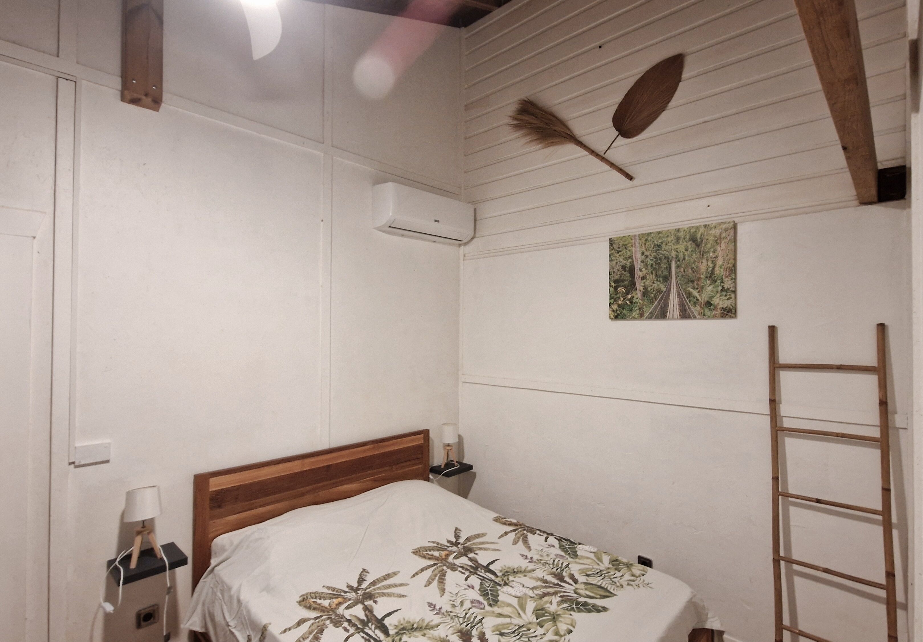Een slaapkamer met een houten hoofdbord, een bed met een bloemenbedekking, een nachtkastje met een lamp, een wandairconditioner, een ladder en een schilderij aan de muur.