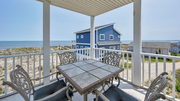 Un espace extérieur abrité, équipé d’une table et de chaises, d’où l’on a vue sur une plage et une maison bleue.