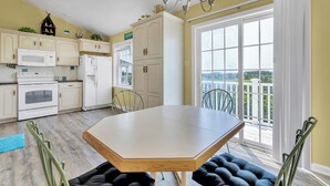 Une cuisine avec des meubles blancs, un four à micro-ondes et un réfrigérateur. On y trouve une table à manger avec des chaises, et une grande fenêtre offre une vue sur l’extérieur.