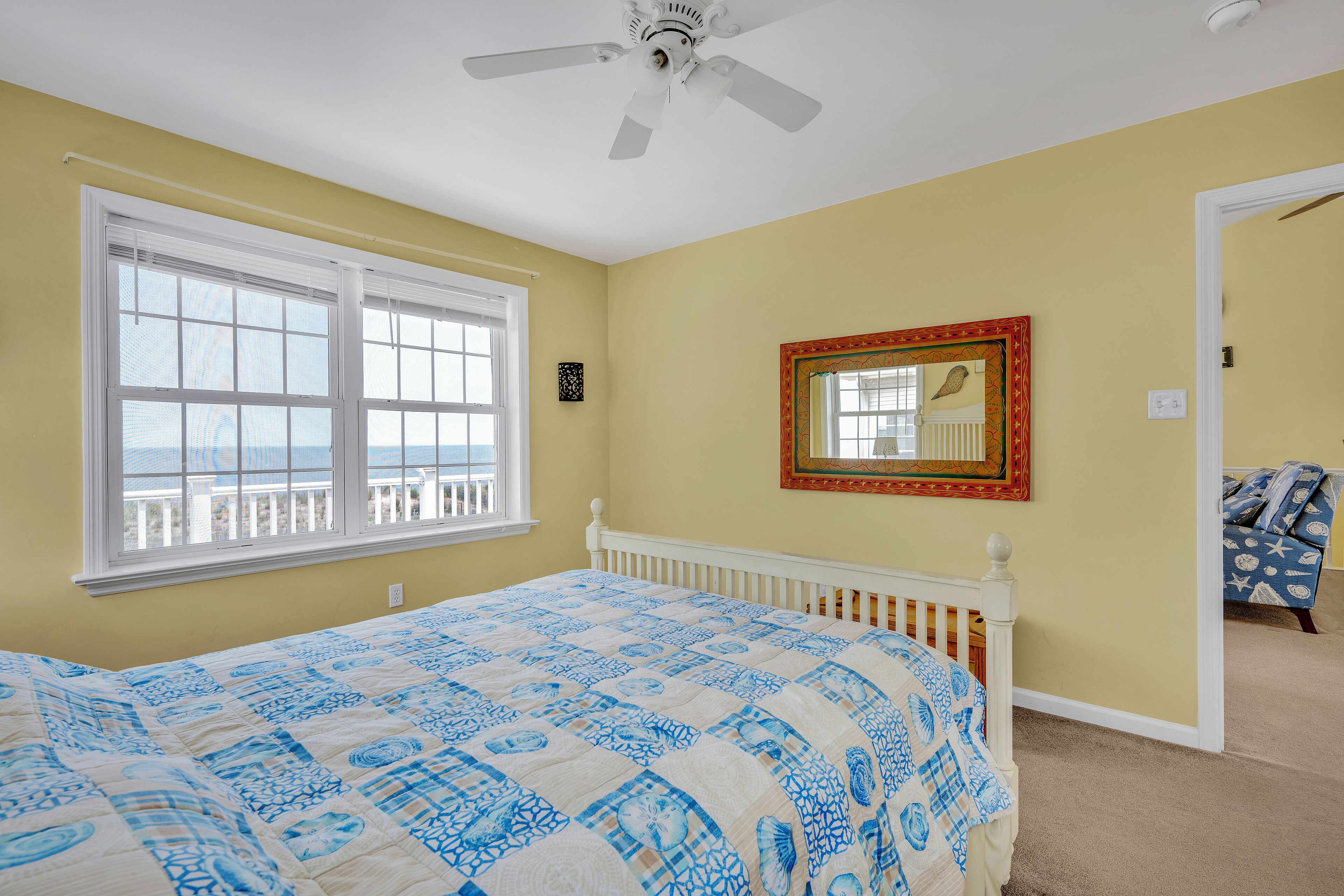 Quarto com cama, ventilador de teto, janela com persianas, espelho e porta para outro cômodo.