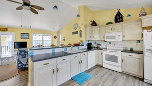 Une cuisine avec des meubles blancs, un four à micro-ondes et une cuisinière. On y trouve également un ventilateur de plafond, un tapis bleu et une fenêtre avec vue.