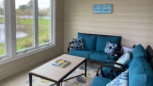 Una habitación con un ventanal grande, un sofá azul con cojines estampados, una mesita con un libro y una pared de madera con un letrero.